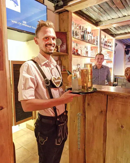 Kellner in Lederhose mit Lebkuchenherz und einem Tablett Bier in der Almstube